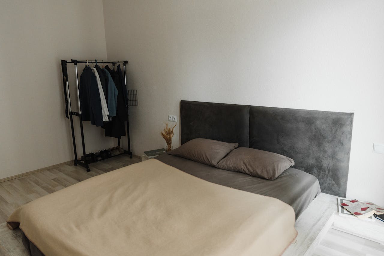 A minimalist bedroom featuring a stylish gray headboard, beige bedding, and a simple clothes rack for a modern look.